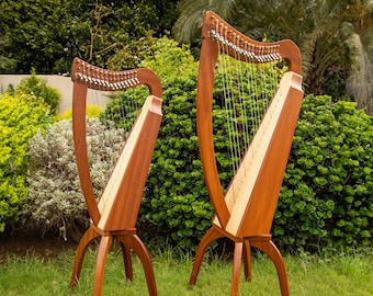 May include: Two wooden harps of different sizes standing on four-legged bases. Each harp features a reddish-brown wooden frame, a light-colored soundboard, and a series of vertical metal strings with tuning pins along the top neck.