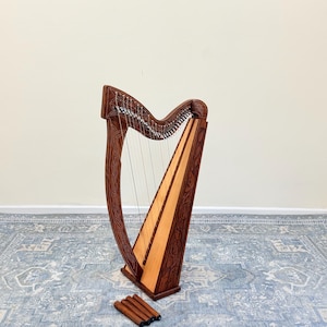 May include: A wooden floor harp featuring a dark brown frame with intricate carved knotwork patterns and a light-colored soundboard. The instrument has numerous vertical strings and is accompanied by four separate, matching dark brown wooden legs.