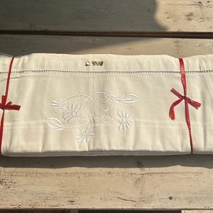 May include: A folded off-white textile piece featuring white floral embroidery in the center and a decorative hemstitch border along the top edge. The fabric is secured with two thin red ribbons tied in bows on the left and right sides.