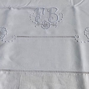 Puede incluir: Mantel de lino blanco con detalles bordados de flores e iniciales. El diseño incluye dos grandes flores bordadas, un monograma central y costuras decorativas en los bordes. Un diseño clásico y elegante.