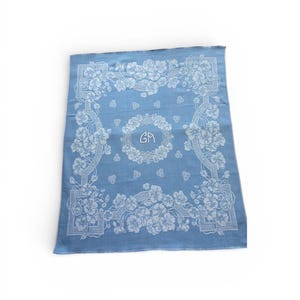 May include: A pale blue bandana with a white floral pattern. The design includes a floral border and a central circular emblem with the initials "GA". The fabric appears to be soft, possibly cotton or a similar material.