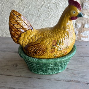 Può includere: Un contenitore in ceramica a forma di gallina nei toni del giallo e del marrone, con cresta rossa e becco verde. La gallina è appoggiata su una base verde a forma di vimini. Il contenitore è probabilmente utilizzato per scopi decorativi o funzionali.