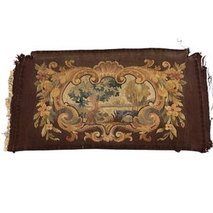 May include: A rectangular brown tapestry featuring a landscape scene within a gold, green, and beige floral and scrollwork frame. The tapestry has a fringed edge and a detailed woven texture, typical of traditional craftsmanship.