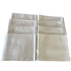 Puede incluir: Un conjunto de servilletas de tela blancas, apiladas en dos montones separados. Las servilletas tienen un sutil diseño floral en relieve. La imagen está bien iluminada, resaltando la textura y el estado impecable de la tela. Ideal para cenas formales.