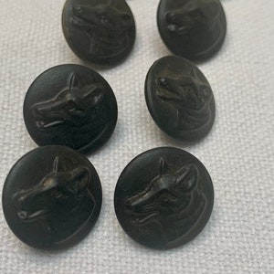 Könnte beinhalten: Acht schwarze Knöpfe mit einem erhabenen Wolfskopf-Design. Die Knöpfe sind rund und haben eine glatte, matte Oberfläche.