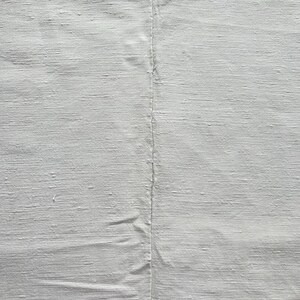 Può includere: Primo piano di un tessuto bianco con una cucitura centrale. Il materiale presenta una trama visibile, forse lino o un tessuto simile. Sono presenti sottili variazioni di tono e consistenza.