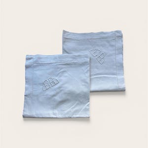 May include: Two light blue pillowcases with embroidered initials. The pillowcases are rectangular and have a soft, slightly wrinkled appearance. Each pillowcase features a stylized monogram in the upper corner, adding a touch of elegance to the bedding.