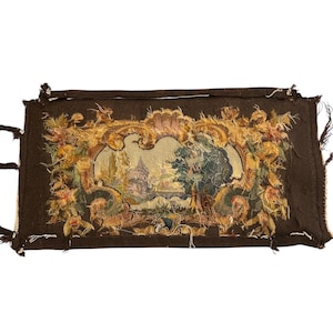 May include: A rectangular tapestry with a dark brown border and a central landscape scene featuring buildings and trees. The scene is framed by ornate gold and floral embellishments. Frayed edges and tassels are visible.