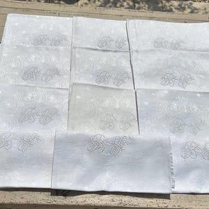 May include: A set of ten white rectangular napkins arranged in a grid. Each napkin features a tonal damask pattern and a central embroidered monogram consisting of the intertwined letters B and S in a cursive script style with floral accents.