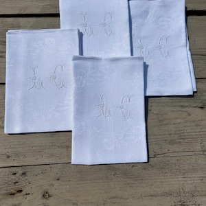 May include: A set of four rectangular white napkins featuring a tonal damask floral pattern and an embroidered monogram consisting of the letters H and C in the center of each piece, arranged in a cluster on a wooden surface.