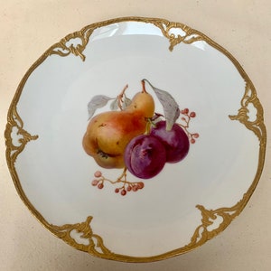 Puede incluir: Un plato de porcelana blanca con un borde dorado y un diseño pintado de una pera y dos ciruelas. El plato está decorado con un patrón floral dorado alrededor del borde.