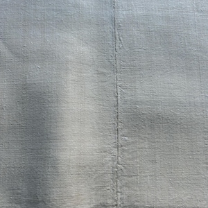 May include: Close-up of a light gray linen fabric with a visible vertical seam. The fabric has a textured appearance, with subtle variations in color and a slightly wrinkled texture.