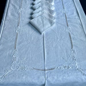 以下が含まれることがあります： 白いリネンのテーブルクロス、刺繍された花のディテールと、お揃いのナプキンのセット。 テーブルクロスは、複雑な透かし彫りと装飾的なボーダーが特徴です。 ナプキンはテーブルにきちんと積み重ねられており、繊細な刺繍が施されています。