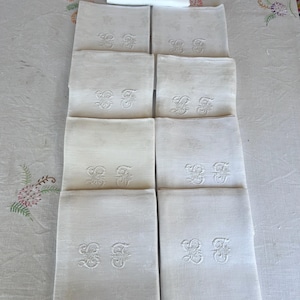 May include: A collection of white linen napkins, each embroidered with an ornate monogram. The napkins are neatly stacked on a white tablecloth, showcasing their crisp, clean appearance. The embroidery adds a touch of elegance.