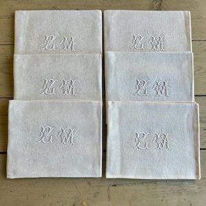 May include: A set of six white linen napkins. Each napkin features an embroidered monogram in a delicate, ornate script. The napkins are neatly folded and arranged, showcasing their crisp, clean appearance. The background is a light-colored wooden surface.
