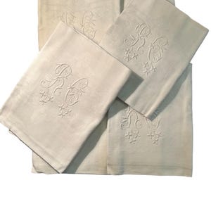 May include: A collection of off-white linen napkins. Each napkin features an embroidered monogram with floral accents. The napkins are arranged in a slightly overlapping manner, showcasing the delicate details of the embroidery and the texture of the fabric.