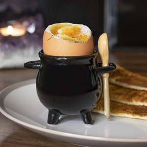 May include: A black ceramic egg cup with a handle and three feet, holding a soft-boiled egg. A small wooden knife is placed on the side. The egg cup is on a white plate with toast.