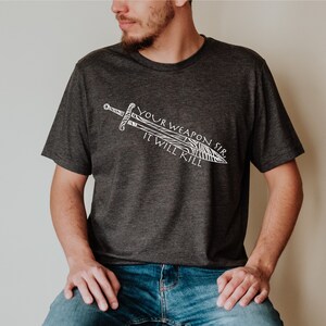 May include: A dark gray t-shirt with a white graphic of a sword and the text "Your Weapon Sir, It Will Kill."