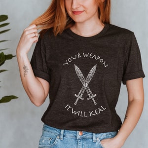 Op de afbeelding: Een donker grijs t-shirt met een witte afbeelding van twee gekruiste zwaarden en de tekst "YOUR WEAPON IT WILL KEAL".