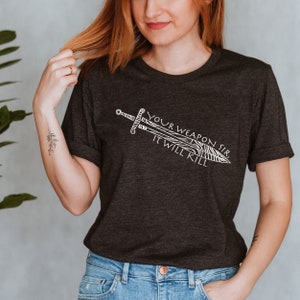 May include: A dark gray t-shirt with a white graphic of a sword and the text "Your Weapon Sir It Will Kill."