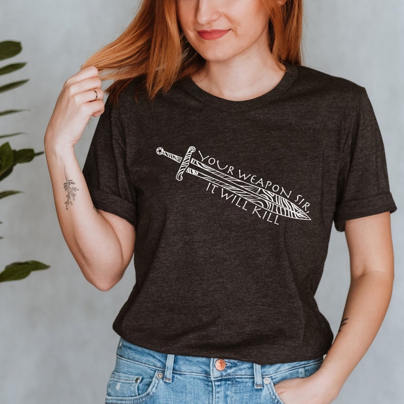 May include: A dark gray t-shirt with a white graphic of a sword and the text "Your Weapon Sir It Will Kill."