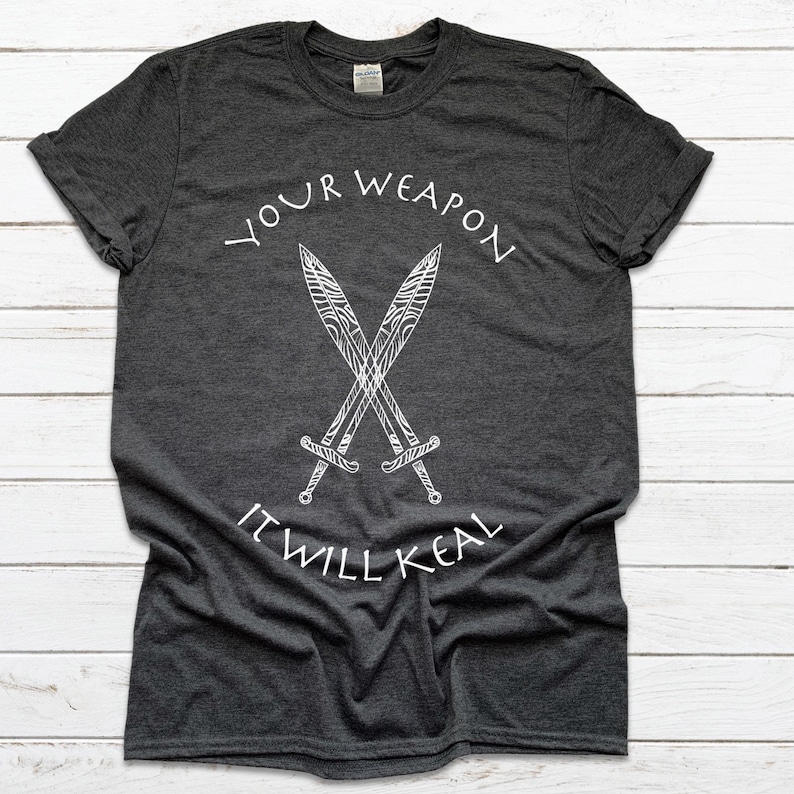 Op de afbeelding: Donkergrijs T-shirt met een witte afbeelding van twee gekruiste zwaarden en de tekst "YOUR WEAPON IT WILL HEAL".