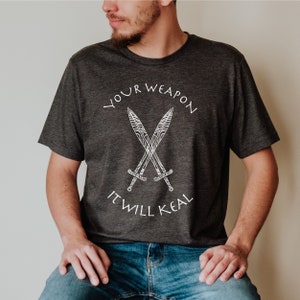 Op de afbeelding: Een donkergrijs T-shirt met een witte afbeelding van twee gekruiste zwaarden en de tekst "YOUR WEAPON IT WILL KEAL".
