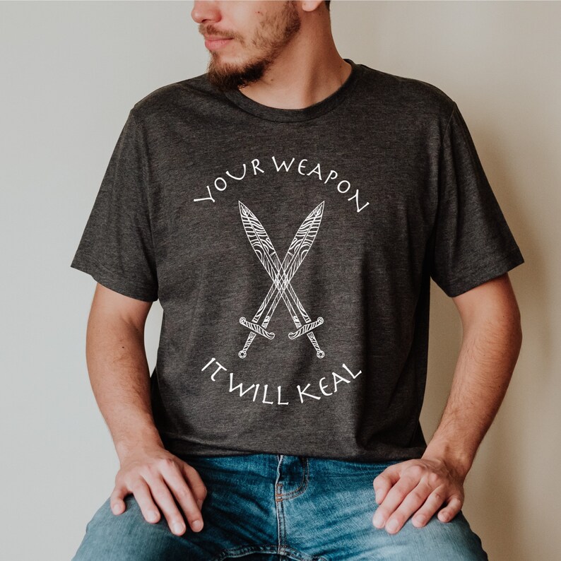 Op de afbeelding: Een donkergrijs T-shirt met een witte afbeelding van twee gekruiste zwaarden en de tekst "YOUR WEAPON IT WILL KEAL".
