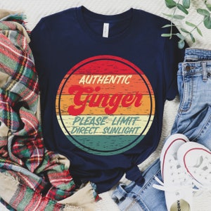 May include: A navy blue t-shirt with a vintage-style graphic of a red, orange, yellow, green, and blue circle with the text "Authentic Ginger Please Limit Direct Sunlight".