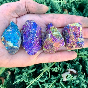 May include: Four iridescent, colorful rocks with shades of blue, purple, and gold are held in a hand. The rocks have a rough, textured surface and vary slightly in shape and size. The background is a blurred view of green foliage.