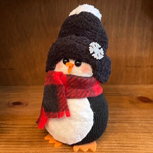 May include: A black and white sock penguin wearing a red and black plaid scarf and a black knit hat with a white pom-pom and a snowflake.