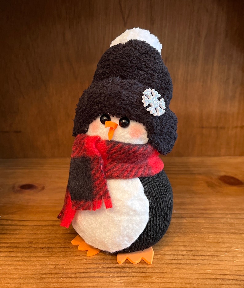 May include: A black and white sock penguin wearing a red and black plaid scarf and a black knit hat with a white pom-pom and a snowflake.