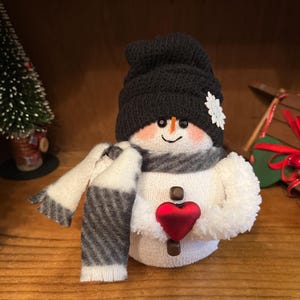 May include: A white snowman wearing a black knitted hat, a white and grey striped scarf, and holding a red heart. The snowman is sitting on a wooden surface.