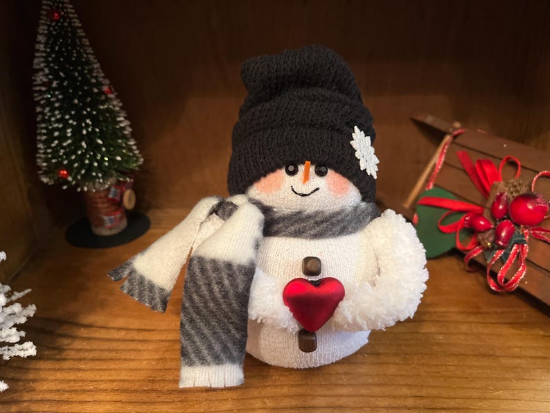 May include: A white snowman wearing a black knitted hat, a white and grey striped scarf, and holding a red heart. The snowman is sitting on a wooden surface.