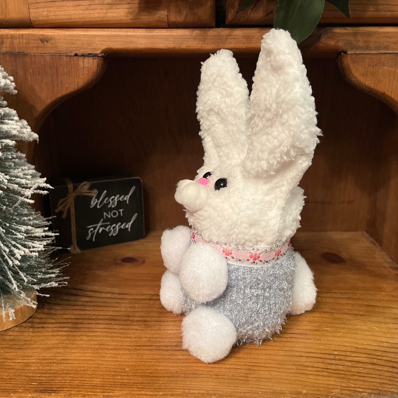 May include: A white bunny plush toy with grey and pink accents. The bunny is sitting on a wooden shelf with a sign that reads "blessed not stressed".