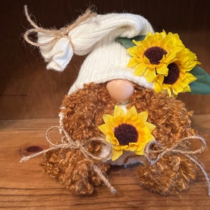May include: A white gnome with brown curly hair wearing a white knit hat and holding three yellow sunflowers.