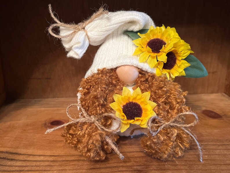 May include: A white gnome with brown curly hair wearing a white knit hat and holding three yellow sunflowers.