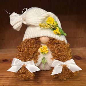 May include: A white gnome with brown curly hair, wearing a white knitted hat with yellow flowers and white bows on the ends of the hair.