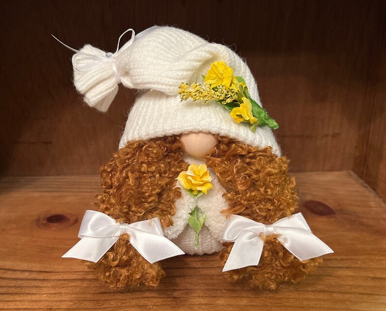 May include: A white gnome with brown curly hair, wearing a white knitted hat with yellow flowers and white bows on the ends of the hair.