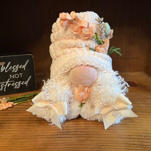 May include: A white gnome with a pink knitted hat and peach-colored flowers. The gnome has a white fluffy beard and white bows on the ends of the beard. The gnome is sitting on a wooden surface.