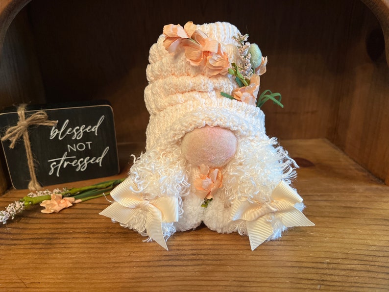 May include: A white gnome with a pink knitted hat and peach-colored flowers. The gnome has a white fluffy beard and white bows on the ends of the beard. The gnome is sitting on a wooden surface.