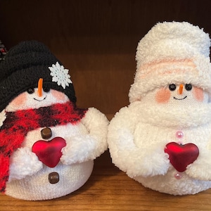 May include: Two handmade sock snowmen holding red felt hearts. One snowman is wearing a black hat, red and black plaid scarf, and white buttons. The other snowman is wearing a white hat and pink buttons.