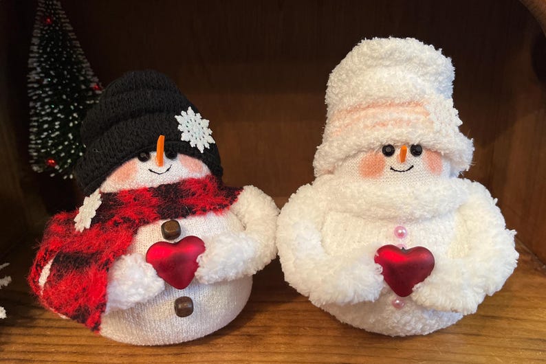 May include: Two handmade sock snowmen holding red felt hearts. One snowman is wearing a black hat, red and black plaid scarf, and white buttons. The other snowman is wearing a white hat and pink buttons.
