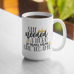Op de afbeelding: Witte keramische koffiemok met een zwart en gouden ontwerp. De mok heeft de tekst "She needed a hero so that's what she became" met een kroon boven het woord "needed".