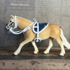 Puede incluir: Un caballo de juguete de color marrón claro que lleva una brida blanca y negra y una silla de montar azul y blanca con ribetes blancos.