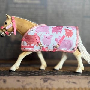 Può includere: Un cavallo giocattolo marrone chiaro che indossa una coperta da cavallo rosa con un motivo a maiali. La coperta ha cinturini rosa e fibbie dorate. Il cavallo ha un guinzaglio rosa con un naso bianco.