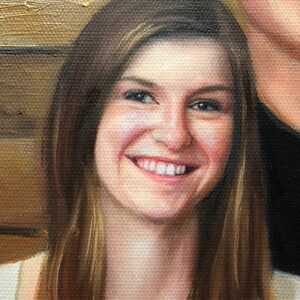 May include: A close-up portrait of a woman with long brown hair smiling. She is wearing a white top and has a warm, friendly expression.