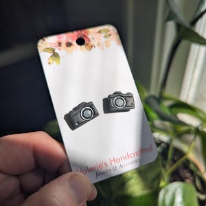May include: A pair of black camera-shaped stud earrings on a white card with floral accents. The card reads "Valerie's Handcrafted Jewelry & Accessories". The earrings are small, detailed, and have a silver-colored lens.