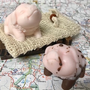 May include: Two pink pig-shaped soaps, one with a smooth surface and the other with brown speckles. The soaps are sitting on a wooden surface with a woven fabric background.