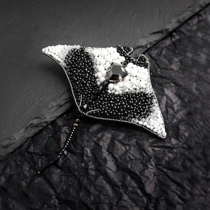 May include: Black and white beaded brooch in the shape of a stingray with a black crystal in the center. The brooch is on a black background.